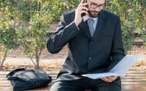 A man, likely a lawyer, wearing a dark pinstripe suit and tie, is sitting on a bench outdoors. He is holding a file or laptop in his lap while talking on a smartphone. A dark satchel is next to him on the bench, suggesting an attorney handling a serious case or taking a professional call while out of the office.