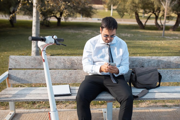 A professional, likely a lawyer, dressed in a white shirt and dark pants, sitting on a wooden bench in a park. He is looking down at and using his smartphone. An electric scooter is parked next to the bench, and a laptop/folder and a satchel are on or near the bench, suggesting a professional taking a work break outdoors.