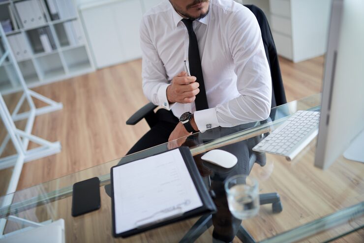 A cropped overhead shot of a person, likely a lawyer, seated at a glass desk in an office. They are wearing a white dress shirt, black tie, and a watch, holding a pen. Documents on a clipboard, a smartphone, a computer mouse, and a glass of water are visible on the desk, suggesting a professional consultation or review of case files.
