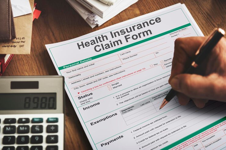 A close-up shot of a hand filling out a Health Insurance Claim Form with a pen, with a calculator showing the number 8980 nearby. The form requires personal details, income status, and exemption information, representing the process of filing an injury claim.