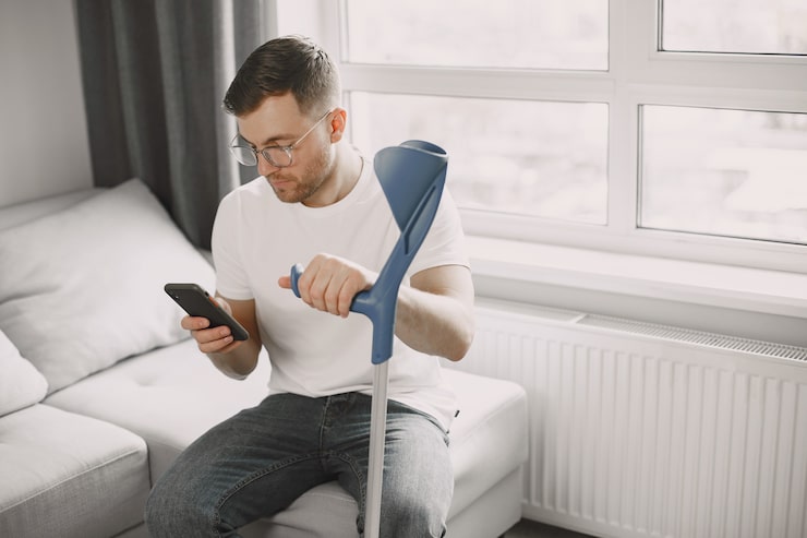 A man with glasses, holding a blue crutch, sits on a couch indoors while looking at his smartphone, suggesting a person with an injury engaging in an activity that could be subject to insurance company surveillance tactics or social media monitoring.