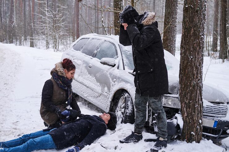 People attending to an injured person lying in the snow next to a car crashed into a tree, representing severe winter car accidents in Canada.