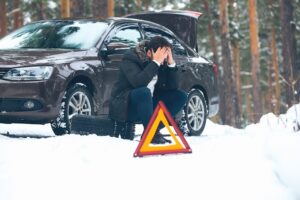 Distressed driver crouches by a snowy car with a flat tire and a warning triangle, illustrating a roadside winter car accident or breakdown.