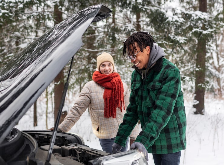 A man and woman look under the raised hood of a car in a snowy forest, depicting a common winter accident like a vehicle breakdown.