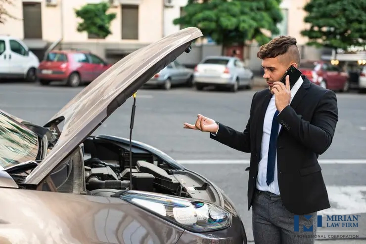 A man in a business suit talks on his phone next to his broken-down car with the hood up.