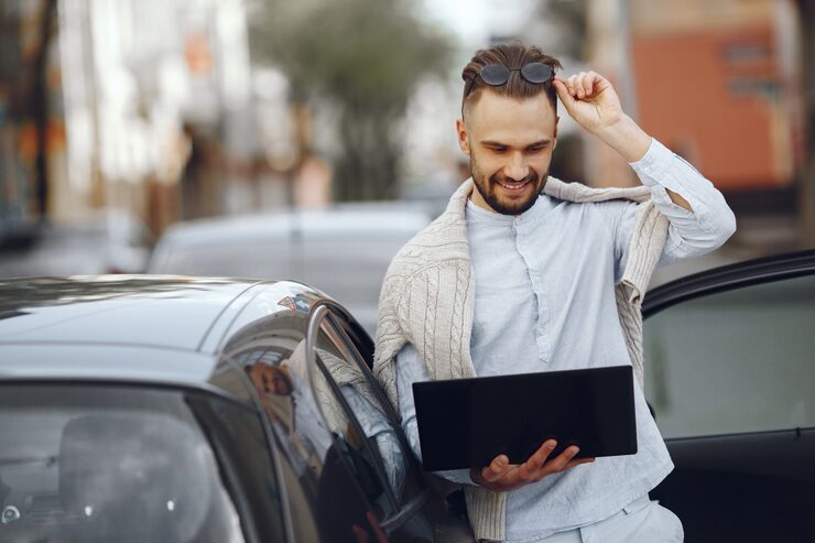 A smiling man in casual wear leans against a car door while looking at a laptop, symbolizing contacting a personal injury lawyer for a free consultation.