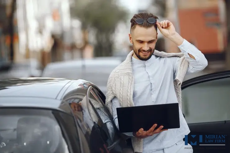 A smiling man in casual wear leans against a car door while looking at a laptop.