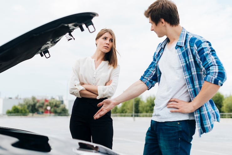 A man points at his car's open hood while a woman with crossed arms looks on, illustrating a post-accident dispute or discussion.