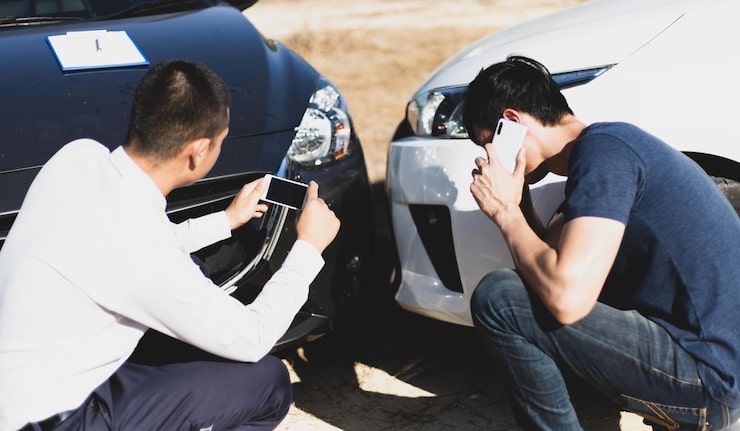 Two drivers examine a minor bumper collision, one taking photos with a phone and the other making a call to report the accident.