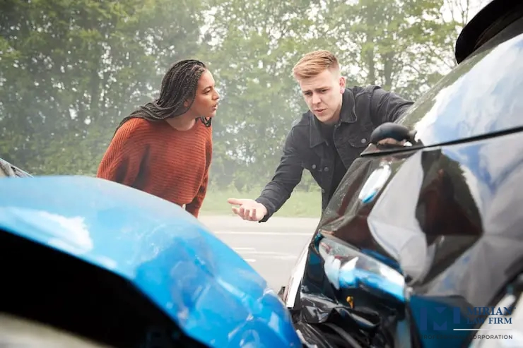 A distressed man and woman stand between two crumpled car bumpers.