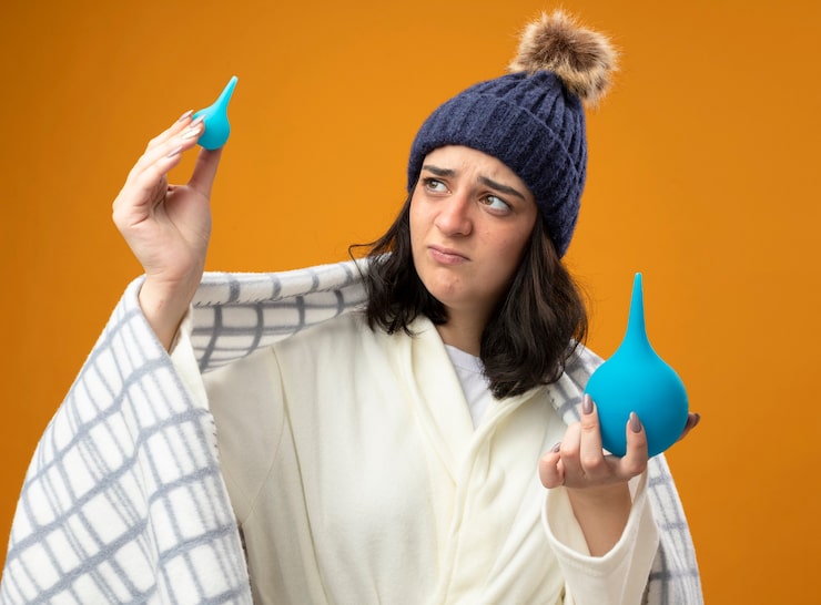 Sick woman wrapped in a blanket, wearing a winter hat, holding two blue medical bulbs with a skeptical look.